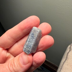 May include: A light blue, raw crystal with a rectangular shape. The crystal has a rough, textured surface and is held in a hand. The crystal's color is a pale, dusty blue.