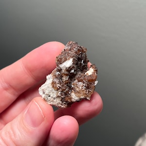 May include: A cluster of brown, cubic crystals with white accents, set on a light-colored matrix. The mineral specimen is held in a hand, showcasing its natural formation and texture. The crystals have a sparkling appearance.