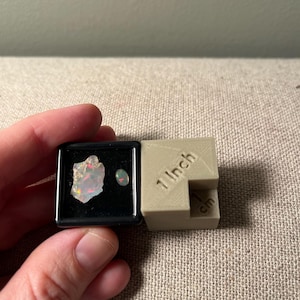 May include: A small black display case holding two opals. One is a rough, multi-colored opal, and the other is a small, round opal. Next to the case is a beige block with "1 Inch" and "cm" printed on it.