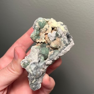 May include: A natural mineral specimen featuring a mix of colors and textures. The rock has a base of gray and white, with clusters of light green and beige formations. The overall shape is irregular and rough, showcasing the raw beauty of the stone.