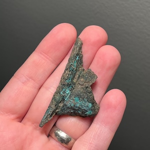 May include: A small, triangular fragment of a dark, textured material with patches of vibrant turquoise-green. The object has a rough, weathered appearance, suggesting age or exposure to the elements. The piece is held in a hand, with a silver ring visible.