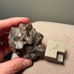 May include: A rough, dark brown mineral specimen with visible crystalline structures and sharp, angular facets. Next to the mineral is a light beige 3D-printed cube with the text 1 Inch clearly embossed on its top surface for scale reference.
