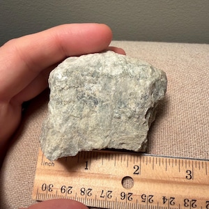May include: A rough, light gray and pale green mineral specimen with a crystalline, textured surface. The rock is positioned against a wooden ruler, showing a width of approximately 2.5 inches across its base, displaying natural mineral formations.