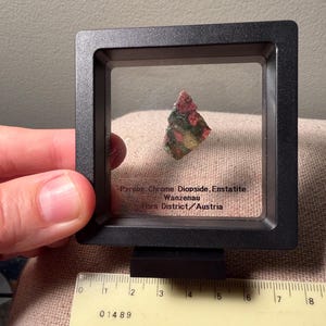 May include: A small, multi-colored mineral specimen containing Pyrope, Chrome Diopside, and Enstatite from Wanzenau, Horn District, Austria, displayed in a square black floating frame. A ruler shows the frame width is approximately 3.5 inches.
