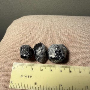 May include: Three polished stones with a black and grey marbled appearance. The stones are positioned above a yellow ruler, showing the largest stone measures approximately 1.5 inches in width and the smallest measures about 0.75 inches.