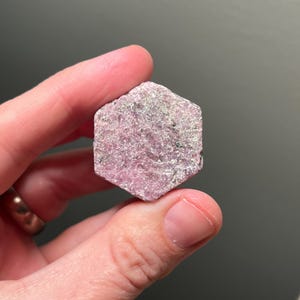 May include: A hexagonal, rough-cut gemstone in shades of pink and gray. The stone is held between two fingers, showcasing its natural, unpolished texture. The gem's surface has a slightly granular appearance, with subtle variations in color.