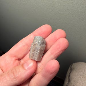 May include: A small, rough-textured, light gray and tan stone with a crystalline appearance. The stone is held in a hand, showcasing its natural, irregular shape. The stone appears to be a mineral specimen.