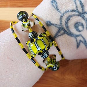 Peut inclure: Un bracelet multi-brins composé de perles en verre jaunes et noires. Le design comprend des perles en verre plus grandes et centrales avec des motifs rayés jaunes, noirs et blancs, alternées avec de petites perles de rocaille sur un fil souple.