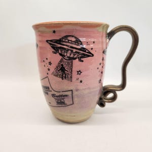 May include: Pink ceramic mug with a black illustration of a UFO, a person being beamed up, and stars. The mug has a unique bronze-colored handle and the word "Butter" is visible on the side.