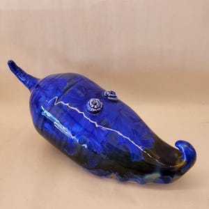 May include: A glossy ceramic sculpture shaped like a chili pepper, featuring a deep cobalt blue glaze with crystalline patterns. The piece has a curved, tapered form with two small, raised circular decorative elements on the upper surface.