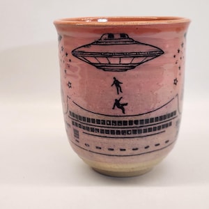 May include: A ceramic mug with a pink and tan gradient. The mug features a black illustration of a UFO, two falling figures, and stars. The design wraps around the mug, creating a unique and artistic look. The mug is suitable for hot or cold beverages.
