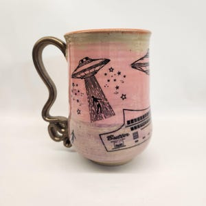 May include: A ceramic mug with a pink and green gradient. The mug features a black illustration of a UFO beaming up a person, with stars and the word "Butter" below. The handle is a bronze color.