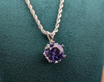 May include: A silver necklace with a twisted rope chain and a pendant featuring a large, round, purple gemstone. The gemstone is set in a silver, pronged setting. The necklace is displayed against a dark green background.