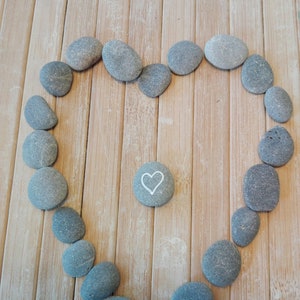 May include: A heart shape made of grey stones on a wooden surface. A single grey stone with a white heart drawn on it is in the centre of the heart shape.