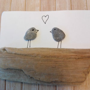 May include: Two grey stone birds with black eyes and legs are perched on a piece of driftwood. A heart is drawn above them on a white background.