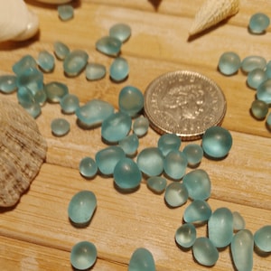 May include: A collection of smooth, light blue sea glass pebbles on a wooden surface. The sea glass is arranged in a cluster, with some pieces overlapping. A silver coin is also visible in the image.