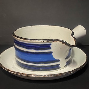 Könnte beinhalten: Eine weiße Keramik-Sauciere mit blauem Streifendesign und kleinem Griff, die auf einer passenden Untertasse steht. Der Rand und die Kanten haben eine braune Umrandung. Das Set ist zum Servieren von Saucen bestimmt.