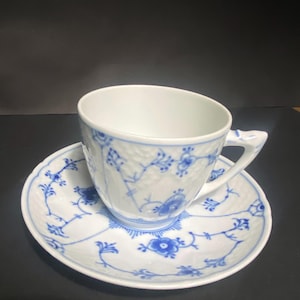 Puede incluir: Una taza de té y un platillo de porcelana blanca con un patrón floral azul. La taza tiene un asa y el platillo tiene un borde festoneado.