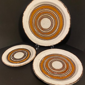 May include: Set of three ceramic plates with a white background and concentric brown and orange circles. The plates are different sizes.