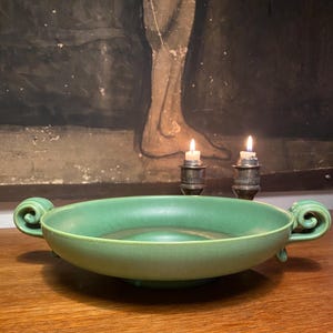 May include: A green ceramic serving bowl with two handles. The bowl has a raised rim and a slightly flared base. The bowl is sitting on a wooden surface.