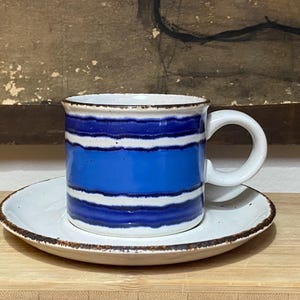 Może przedstawiać: Ceramiczny zestaw filiżanki i spodka do kawy. Filiżanka ma poziome niebieskie i białe paski z białym uchwytem. Spodek jest biały z brązową obwódką. Zestaw stoi na drewnianej powierzchni.