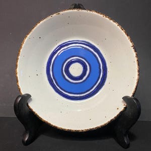 Könnte beinhalten: Eine Keramikschale mit weißem Innenraum und einem blauen Design aus konzentrischen Kreisen in der Mitte. Der Rand hat einen braunen, strukturierten Rand. Die Schale wird auf einem dunklen Holzständer präsentiert, der ihr Design hervorhebt.
