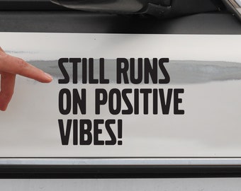 May include: A white car with a black sticker that reads "Still Runs On Positive Vibes!"
