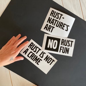 Pode incluir: Três adesivos brancos com texto preto em uma superfície preta. Os adesivos dizem "Rust is not a crime", "No Rust Fun" e "Rust - Nature's Art".