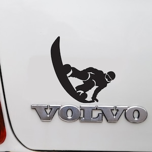 Puede incluir: Volvo blanco familiar con una pegatina de snowboarder negro en la parte trasera. El coche tiene un parachoques negro y una moldura cromada.