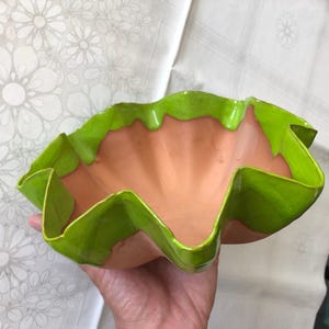 Könnte beinhalten: Eine Keramikschale mit einem gewellten, wellenförmigen Rand, der in einem leuchtenden Limettengrün glasiert ist. Das Innere und die äußere Basis der Schale haben ein natürliches, unglasiertes Terrakotta-Finish, das mit dem glänzenden grünen Rand kontrastiert.
