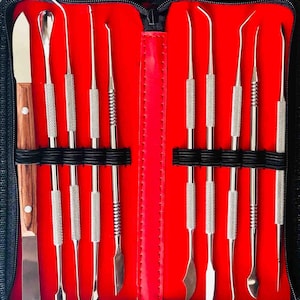 Puede incluir: Un conjunto de 10 herramientas de escultura de acero inoxidable en una funda negra con cremallera y un interior rojo. Las herramientas están dispuestas en filas e incluyen una variedad de formas y tamaños.