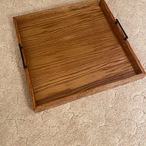 May include: A square wooden serving tray featuring a warm brown wood grain finish and raised edges. The tray is equipped with two slim, matte black metal handles attached to opposite sides, providing a functional grip for carrying items.
