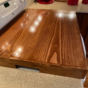 May include: A rectangular, stained wooden stove cover in a warm brown hue. The cover is designed to fit over a standard stove top, providing additional counter space. The wood grain is visible, adding to the rustic aesthetic. The cover has a cutout for the stove controls.