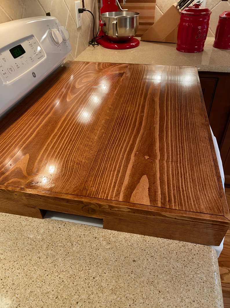 May include: A rectangular, stained wooden stove cover in a warm brown hue. The cover is designed to fit over a standard stove top, providing additional counter space. The wood grain is visible, adding to the rustic aesthetic. The cover has a cutout for the stove controls.