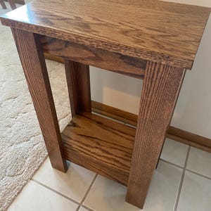 May include: A rectangular wooden side table with a medium brown finish and visible wood grain. The table features a flat top surface, four sturdy vertical legs, and a lower shelf positioned between the legs, providing an open storage area.