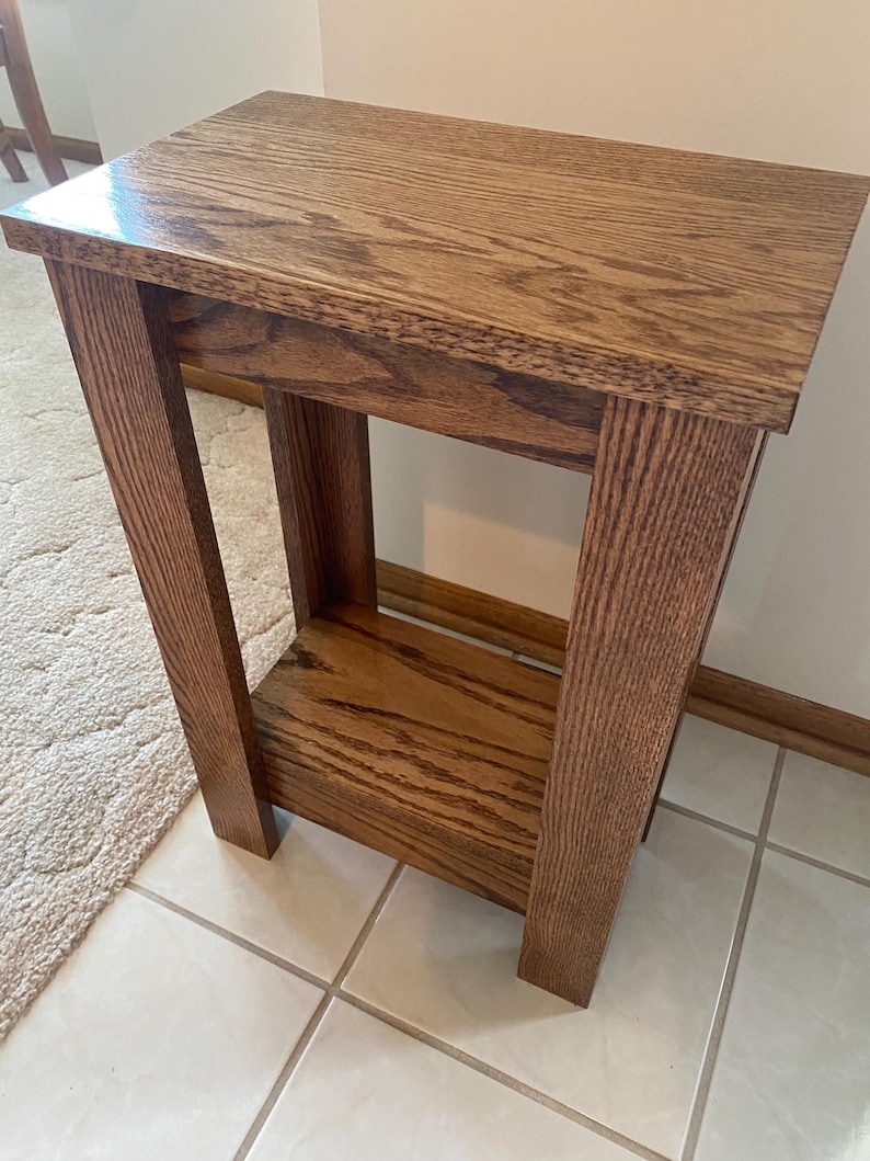 May include: A rectangular wooden side table with a medium brown finish and visible wood grain. The table features a flat top surface, four sturdy vertical legs, and a lower shelf positioned between the legs, providing an open storage area.