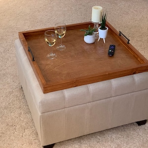 May include: A wooden tray with black handles sits on top of a beige ottoman. The tray has two wine glasses, a small potted plant, a candle, and a remote control on it.