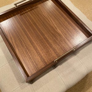 May include: A rectangular wooden serving tray with a rich brown finish and brass-colored handles. The tray has a smooth, polished surface and a raised edge. The handles are attached to the shorter sides of the tray. The tray is resting on a beige upholstered ottoman.