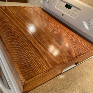 May include: A stained wooden stove top cover in a warm brown hue. The cover is rectangular and fits over a white electric stove. The wood grain is visible, and the surface is smooth and glossy.