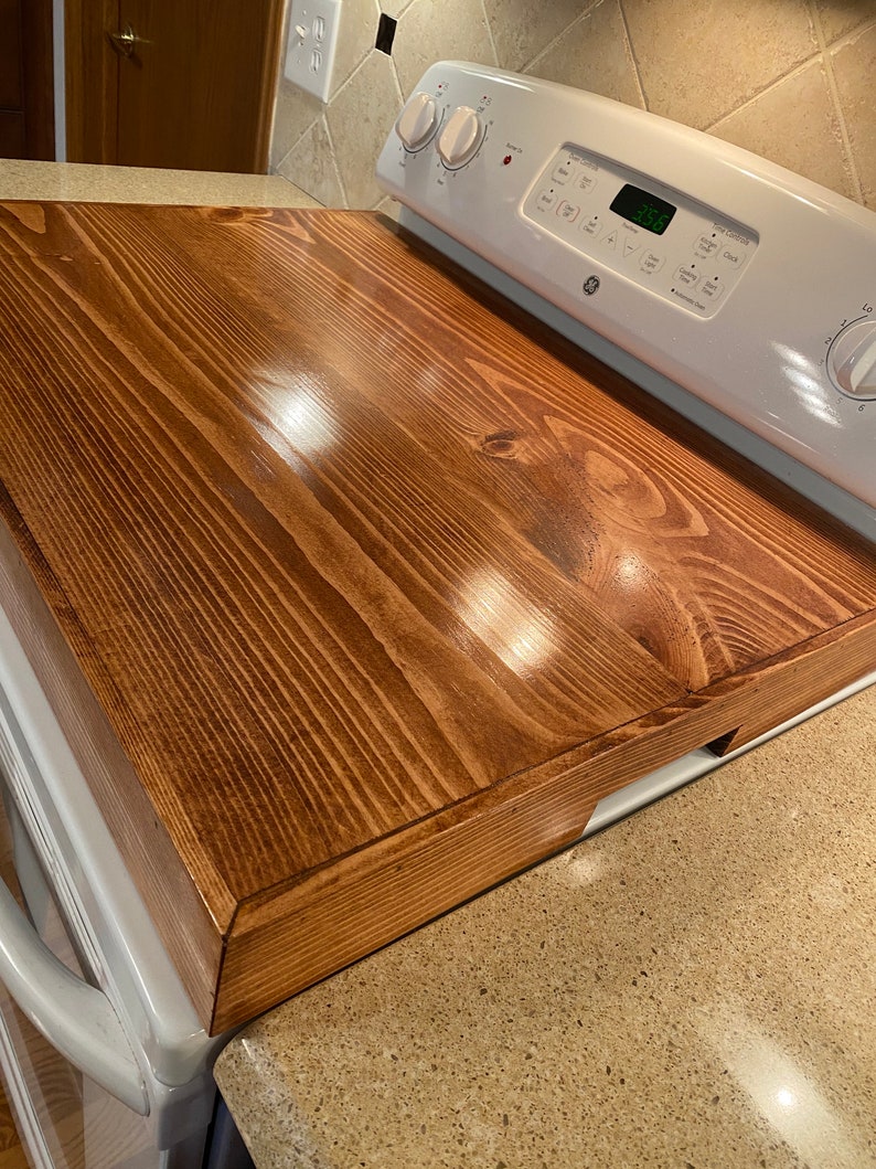 May include: A stained wooden stove top cover in a warm brown hue. The cover is rectangular and fits over a white electric stove. The wood grain is visible, and the surface is smooth and glossy.