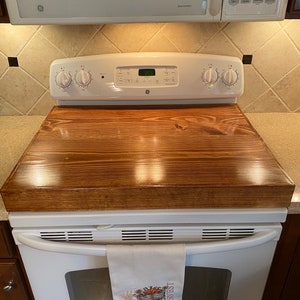 May include: A wooden stove top cover in a rich brown stain, resting on a white GE stove. A white towel with an embroidered autumn harvest design hangs from the oven handle. A microwave is above the stove.