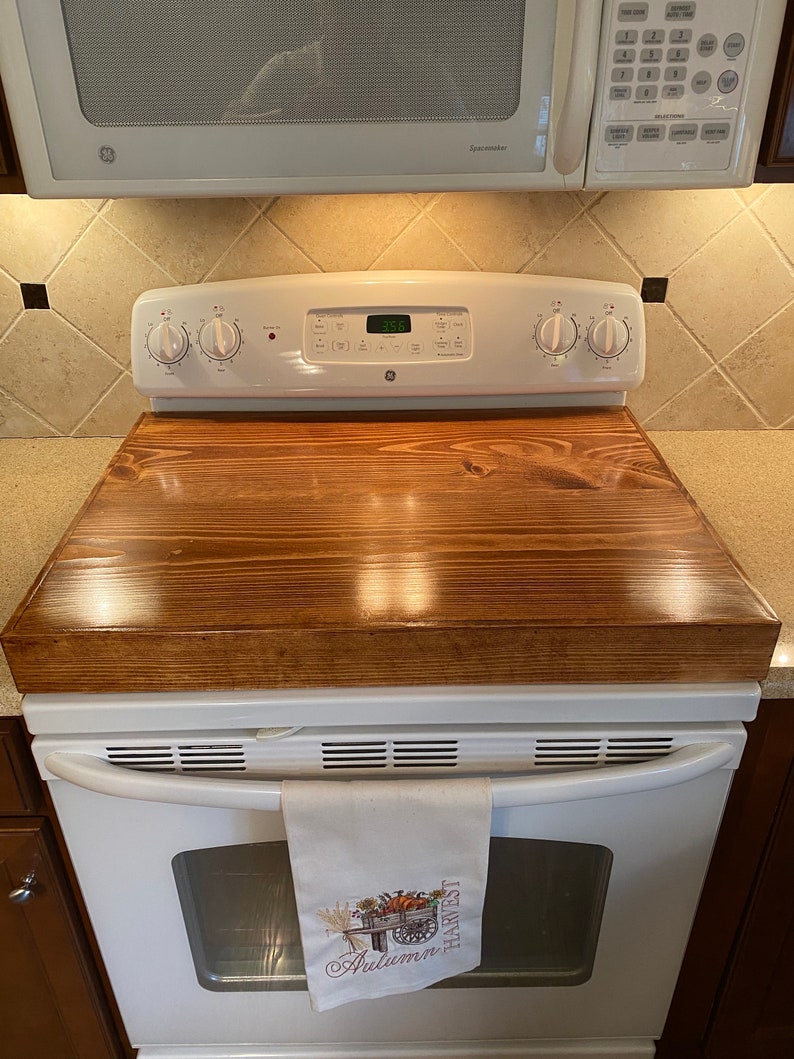 May include: A wooden stove top cover in a rich brown stain, resting on a white GE stove. A white towel with an embroidered autumn harvest design hangs from the oven handle. A microwave is above the stove.