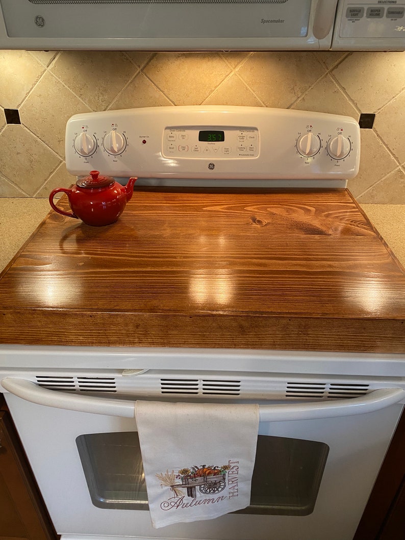 May include: A wooden stove top cover with a rich brown finish sits atop a white stove. A small red teapot rests on the cover. A white oven door with a towel that reads "Autumn Harvest" hangs from the handle.