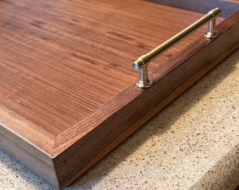 May include: A rectangular wooden serving tray featuring a rich brown wood grain finish and a single polished brass metal handle attached to the side frame. The tray has raised edges and a flat base, showcasing the natural texture of the wood.