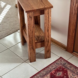 May include: A tall, narrow wooden side table with a rectangular top and a lower shelf. The table is constructed from medium-brown wood with a visible grain pattern, featuring four sturdy, straight legs and a clean, functional design.