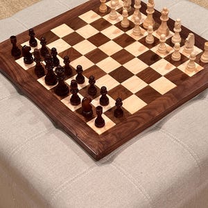 May include: A wooden chess set with a dark brown frame and a checkered playing surface. The chess pieces are arranged on the board, with dark brown and light brown pieces facing each other. The board is set on a light gray upholstered ottoman.