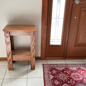 May include: A rectangular wooden side table with a medium brown finish and visible wood grain. The table features a flat top surface, four straight legs, and a lower shelf positioned between the legs, constructed with a simple, open frame design.