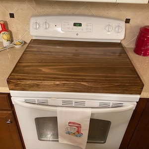 May include: A dark stained wooden stove top cover rests on a white GE electric range. A white kitchen towel with a red mixer design hangs from the oven handle. A microwave is above the stove.