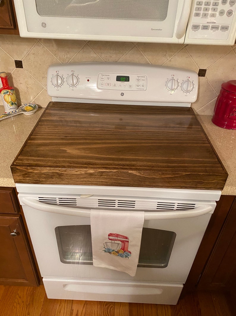 May include: A dark stained wooden stove top cover rests on a white GE electric range. A white kitchen towel with a red mixer design hangs from the oven handle. A microwave is above the stove.