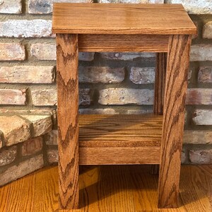 May include: A rectangular wooden side table featuring a medium brown finish with a visible wood grain pattern. The table has a flat top surface, four straight legs, and a lower shelf positioned between the legs, constructed from solid wood.